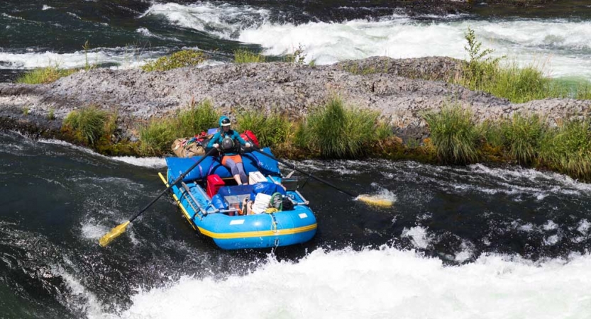 A person paddles a packed raft through whitewater in front of a rocky shore.